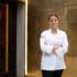 A cook stands in front of a restaurant in the snow and smiles at the camera.