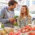 A couple is standing in front of a stall at the Viktualienmarkt in Munich. A man is holding a bunch of asparagus in his hands.