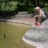 A woman in a baseball cap and tank top sits on the shore of a small lake in which a duck is swimming.