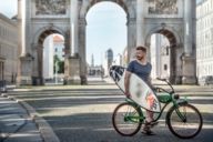 A male surfer with his surfboard and bicycle at the Siegestor in Munich.