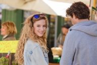 A woman with long curly hair is smiling in front of a stall at the Viktualienmarkt in Munich.