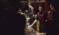 View of a full bar, bartenders dressed in black with red braces work behind the counter.