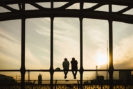 Two people are sitting on the bridge railing of the Hackerbrücke in Munich at sunset.