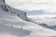 Two skiers skiing down in deep snow in front of a mountain panorama.