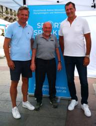Olympic champion Klaus Wolfermann and two other men stand in front of a poster.