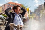 A man is sitting in a street cafe at Gärtnerplatz in Munich. His hands are crossed behind his head.