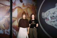 Photograph of two women standing in front of a work of art showing rolled-up slices of ham on the left and the drawn snout of a wild boar on the right.