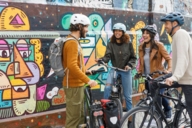 Four cyclists wearing helmets stand in front of a wall painted with graffiti.