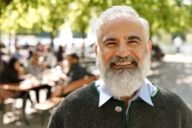 A man with white beard is standing in a beer garden in Munich and is laughing.