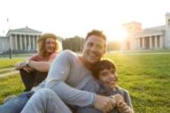 Two men and a boy are sitting on the green grass of the Königsplatz in Munich on a sunny day.