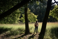 Young woman stands between two trees and looks at a sun-drenched meadow in Munich.