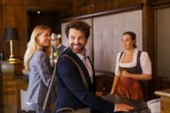 A woman dressed in a dirndl is welcoming a woman and a man in a hotel in Munich.