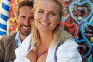 A woman is wearing a dirndl and is smiling in front of gingerbread hearts at the Oktoberfest in Munich. Behind her is sitting a man.