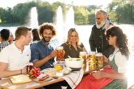 A group of humans is sitting on benches in a beer garden in Munich. On a table between them is traditional food and beer.