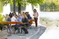 A woman is standing besides a beer bench in a beer garden in Munich. In the background is a lake.