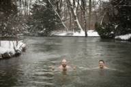 Dwóch mężczyzn pływa zimą w Eisbach w Monachium. Na brzegu leży śnieg.