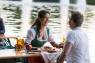 A women and and a man are sitting on benches in a beer garden and are talking and laughing. In the background is a lake.