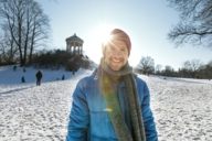 A man is wearing a red knitted hat on a sunny winter day in front of the Monopteros in the English Garden in Munich.