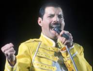 Freddie Mercury sings at the microphone and wears a yellow outfit.