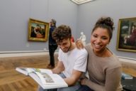 A couple is sitting on stools at Neue Pinakothek in Munich. The man is leafing through a book.