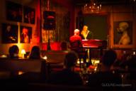 View into a dimly lit bar, in the back a small stage where a man sits at the piano and a woman sings.