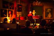 View into a dimly lit bar, in the back a small stage where a man sits at the piano and a woman sings.
