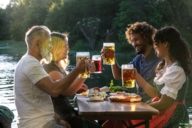 A group of humans is sitting on beer benches in the evening sun with traditional food and beer in a beer garden in Munich.