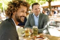 Two men are sitting on benches in a beer garden in Munich. On the table between them are beer mugs and pretzels.