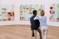 A man is sitting on a stool and a woman stands besides him. They are looking at paintings in the museum Brandhorst in Munich.