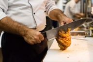 The chef of the restaurant Schneider Bräuhauses München is cutting a knuckle of pork with a big knife.