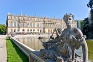 Vue sur la fontaine et la façade du château de Herrenchiemsee. Une statue de pierre repose sur le bord de la fontaine.