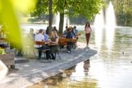 Varias personas están sentadas en bancos en un jardín cervecero de Múnich, junto a un lago, y hablan entre sí. En primer plano hay una hoja verde.