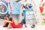 A man is bouldering in the Boulderwelt in Munich and a woman and a man are sitting on the ground and watching the climber.