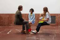 A man and two women, one of whom has a small child on her lap, in a sitting position but without a seat in a room lined with red granite.