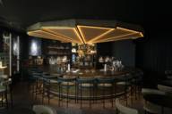 Stylish circular counter with bar stools, in background shelves with drinks, oranges, lemons ,limes and umbrella shaped lighting above the counter in hotel bar Ory in Munich.