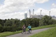 Two women ride their bicycles along a path on the Olympiaberg in Munich. The Olympiastadion is in the background.