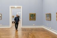 Un uomo con gli occhiali e la barba attraversa la Neue Pinakothek di Monaco di Baviera e guarda i dipinti.