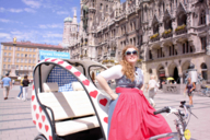 A woman in a dirndl leans against a rickshaw decorated with hearts on Munich's Marienplatz.