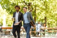 Two men are going through a beer garden in Munich. The man with curly hair is carrying a beer mug and a pretzel.