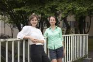 Two women lean against a bridge railing and smile into the camera.