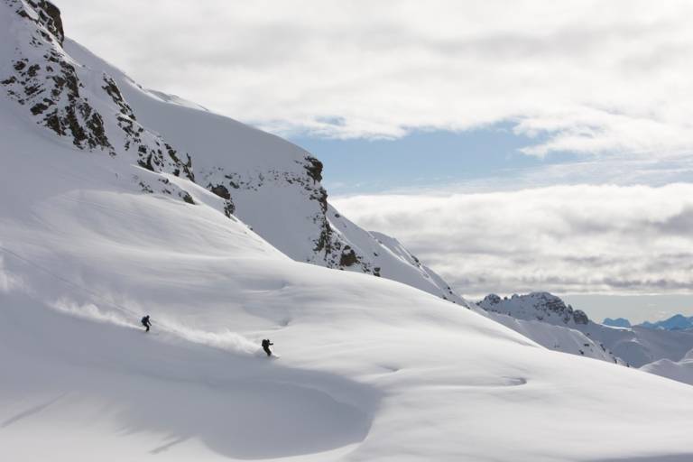 Two skiers skiing down in deep snow in front of a mountain panorama.