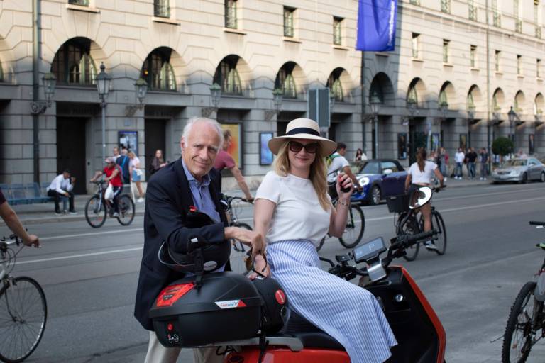 A man and a woman wearing sunglasses and a hat lean against a red scooter on a street in Munich