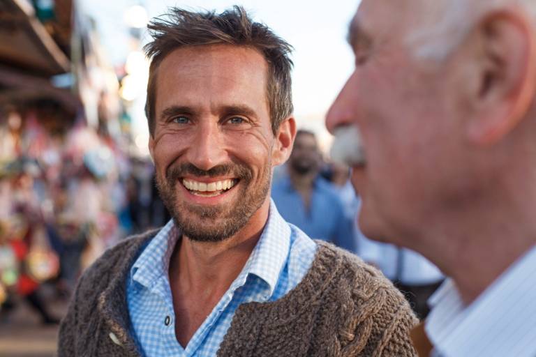 A man is wearing a lederhosen and is smiling at the Oktoberfest in Munich. A man with white hair is standing besides him.