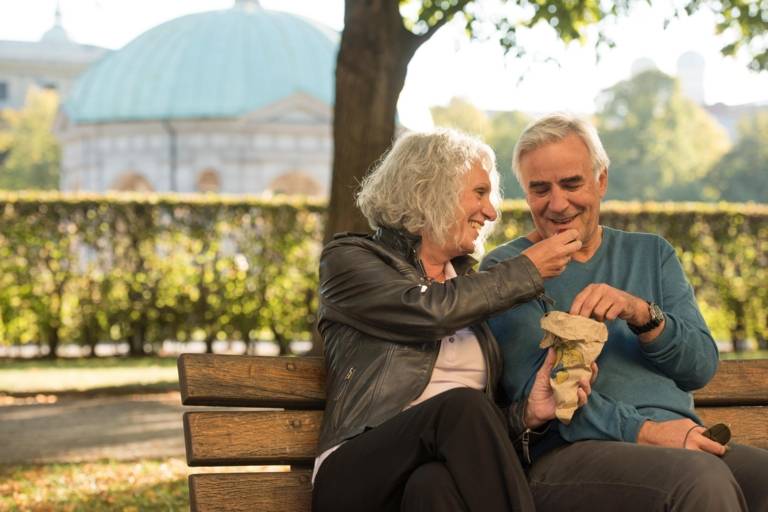 A woman and a man with grey hair are sitting on a bench and eating chestnuts at Hofgarten in Munich.