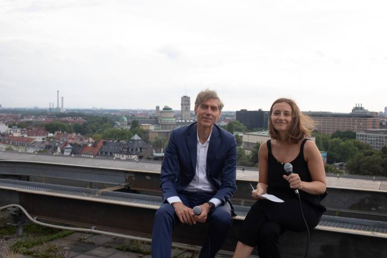 A man and a woman sit with microphones on a roof in Munich.