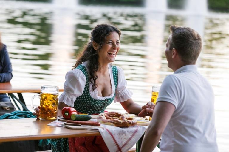 A women and and a man are sitting on benches in a beer garden in Munich and are talking and laughing. In the background there is a lake.