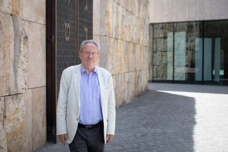 An older man in a jacket stands on a square and looks into the camera.