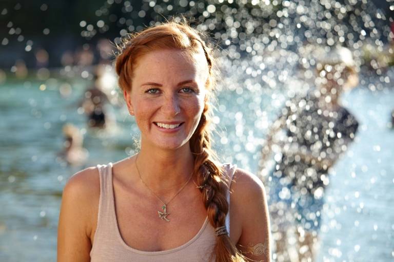 A woman with red long hair is standing and smiling at the Isar River in Munich on a sunny day.