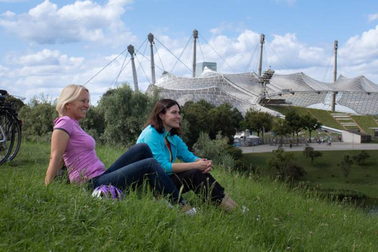 Two women sit on the Olympiaberg in Munich in summer. In the background, bicycles and the Olympiastadion.