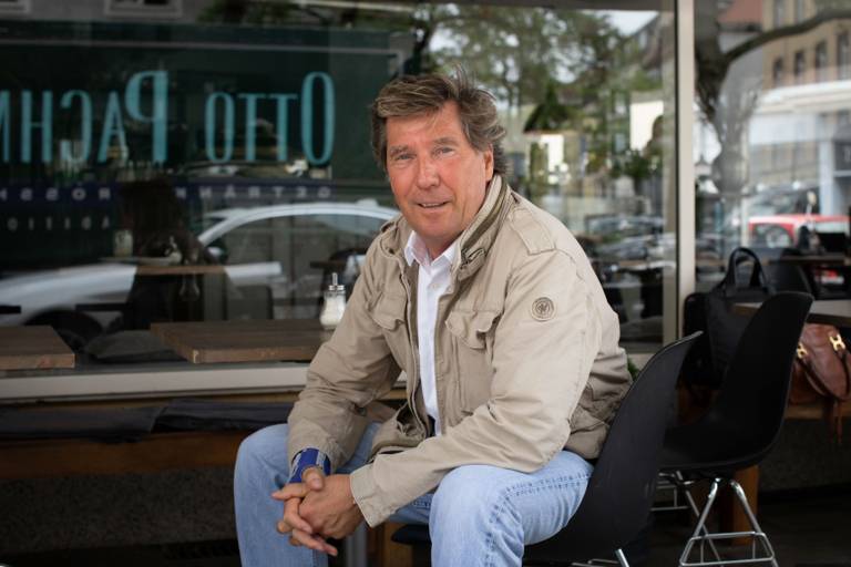 A man sits in front of a café and smiles into the camera.